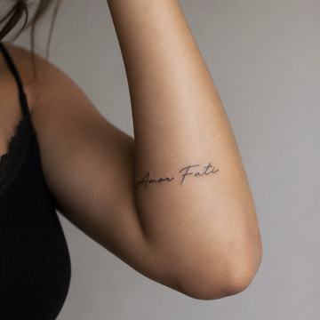 Unterarm einer Person mit kleinem, minimalistischen, kursiven „Amor Fati“-Tattoo in schwarzer Tinte an der Innenseite nahe dem Ellenbogen.