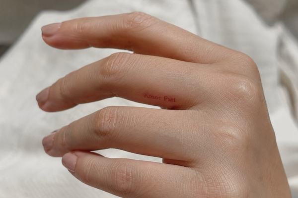 Nahaufnahme einer Hand mit kleinem, minimalistischen „Amor Fati“-Tattoo seitlich am Finger
