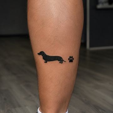 Kleine schwarze Dackel‑Silhouette mit kleinem Pfotenabdruck als minimalistisches Tattoo auf der Wade.