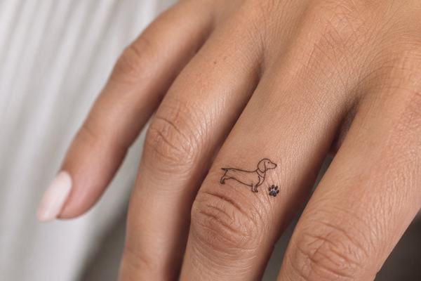 Nahaufnahme einer Hand mit einem kleinen Dackel‑Tattoo im minimalistischen Fineline‑Stil auf dem Finger