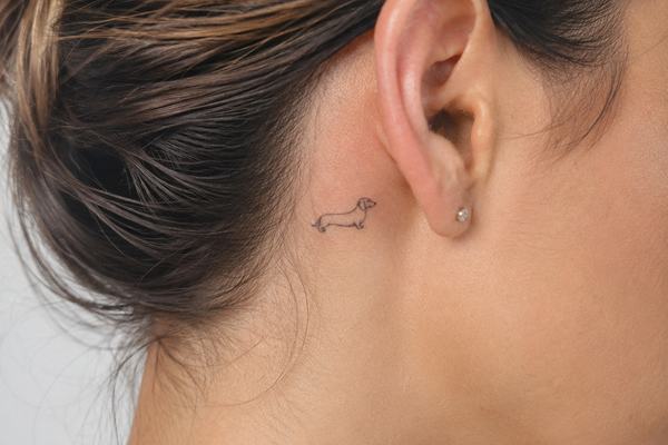 Kleines Dackel‑Tattoo im minimalistischen Fineline‑Stil hinter dem Ohr