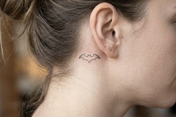 Nahaufnahme eines feinen Batman‑Fledermauszeichens als kleines Schwarz‑Grau‑Tattoo hinter dem Ohr 