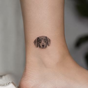 Kleines realistisches Dackelkopf‑Tattoo in feinem Schwarz‑Grau am inneren Knöchel – dezentes Fineline‑Motiv, ideal für kleine, persönliche Dackel‑Designs.